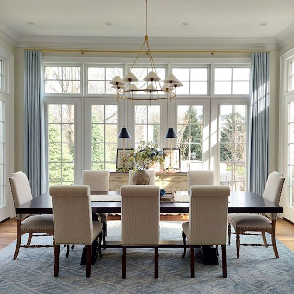 Dining room with a long dark table, cream chairs, blue rug, and large windows with light blue curtains.