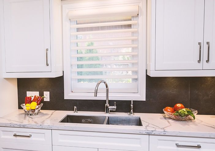 White kitchen sink with window above, marble countertops, and cabinets. Bowls of fruit.