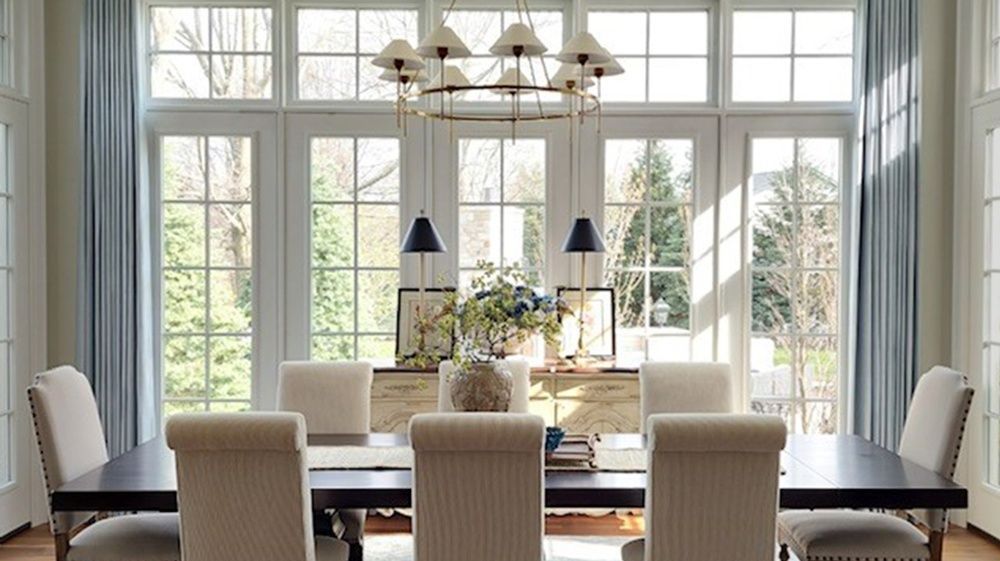 Dining room with large windows, dark table, beige chairs, light blue curtains, and a chandelier.