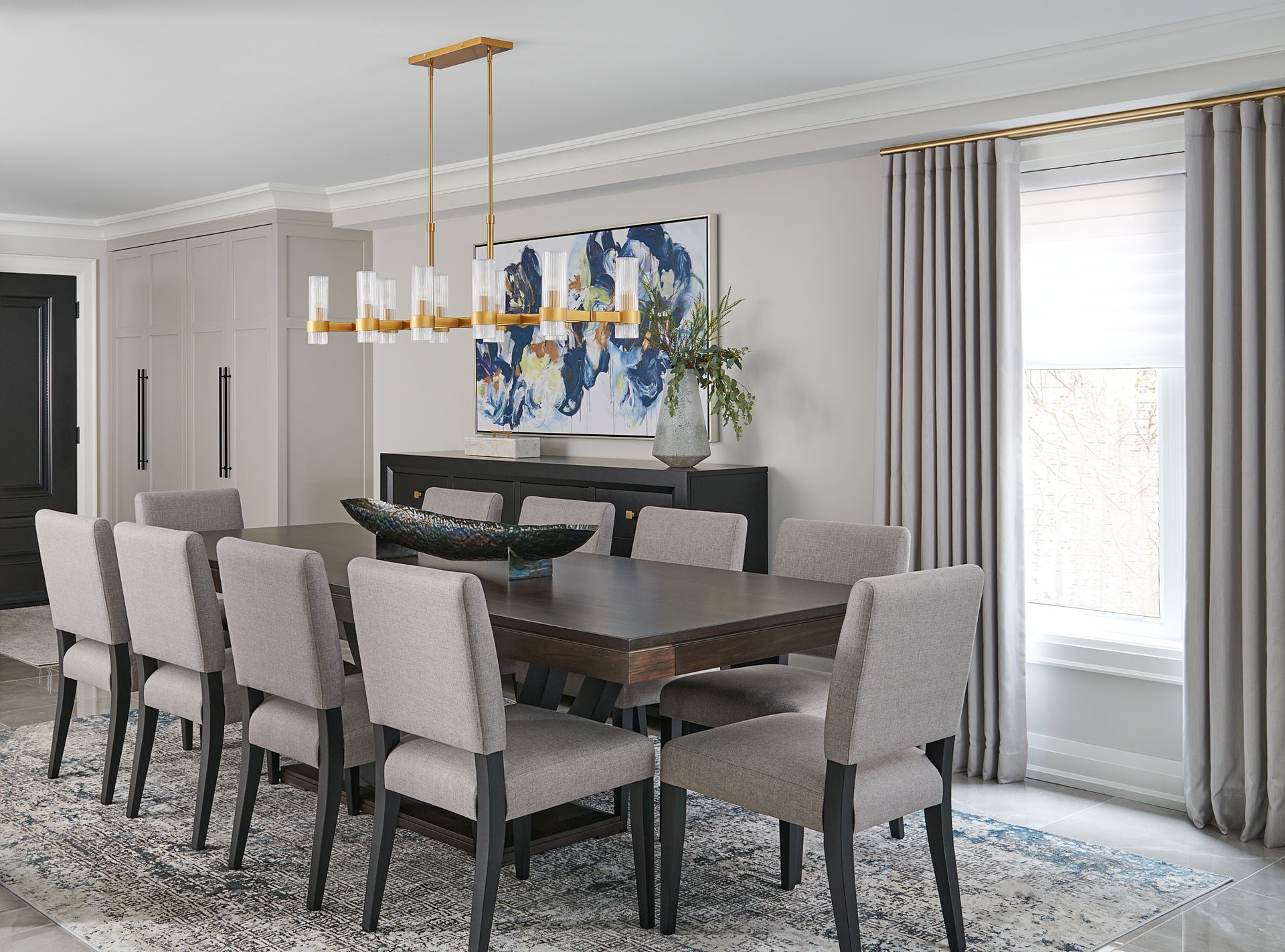 Dining room with long wooden table, ten chairs, and chandelier. Gray curtains, artwork, and cabinet.