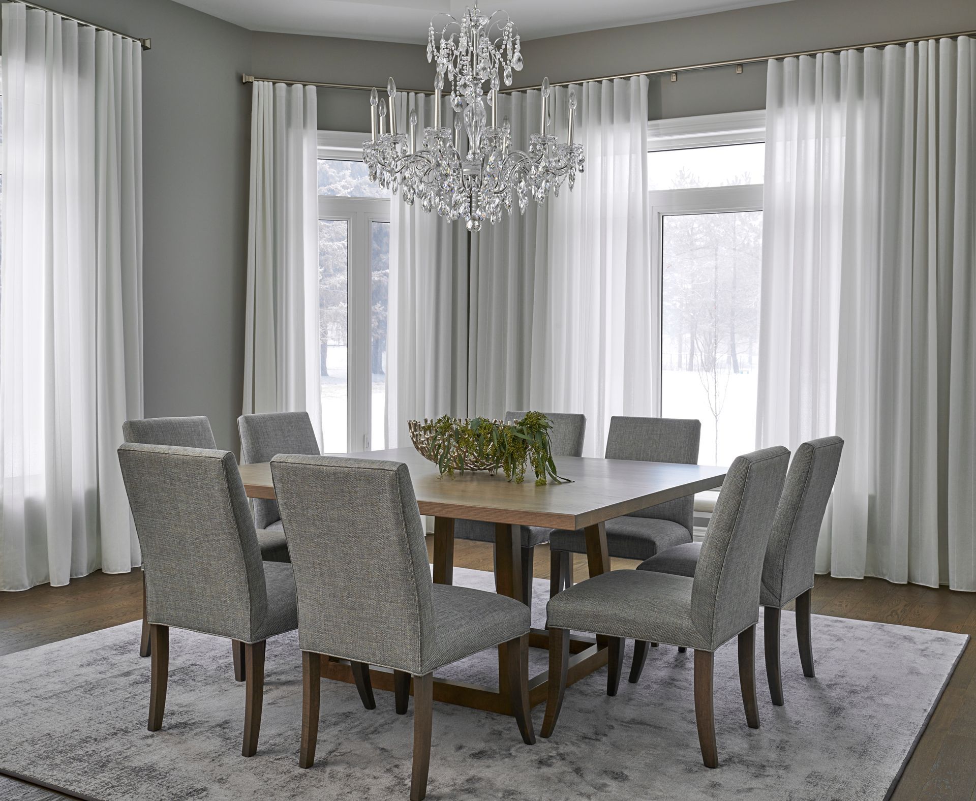 Dining room with a wooden table, gray upholstered chairs, and white curtains. A chandelier hangs above.
