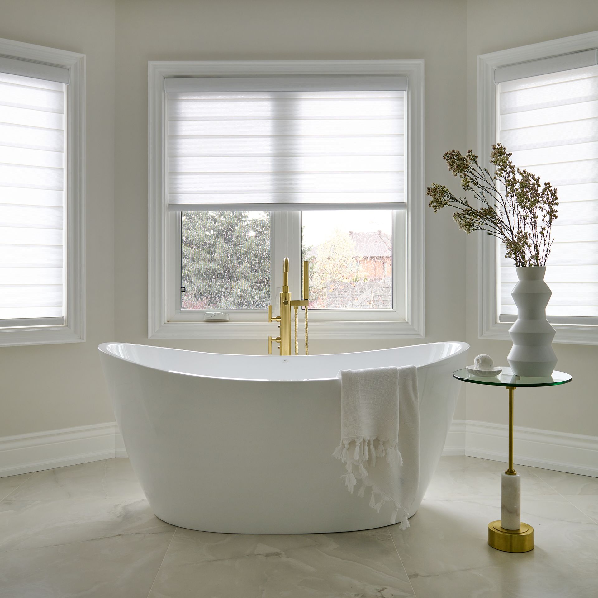 White oval bathtub in a bright bathroom with windows, gold faucet, and a side table with decor.