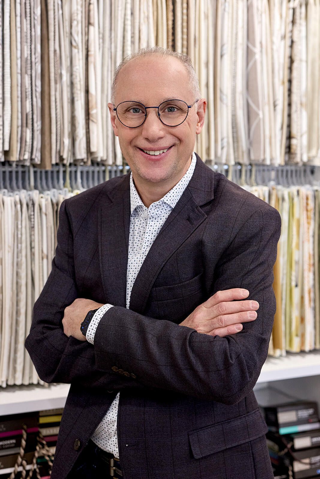 Man in glasses and blazer smiles, arms crossed, in front of fabric samples.