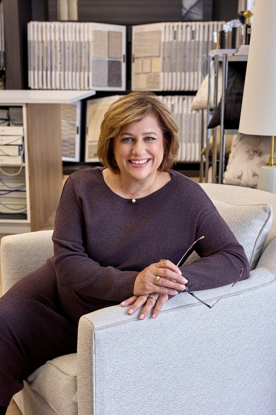 Woman in a purple sweater seated in a light gray armchair, smiling. She holds glasses, in a home decor store.
