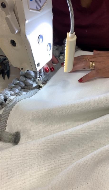 Sewing machine stitching white fabric, with attached gray trim. Person's hands guide the material.