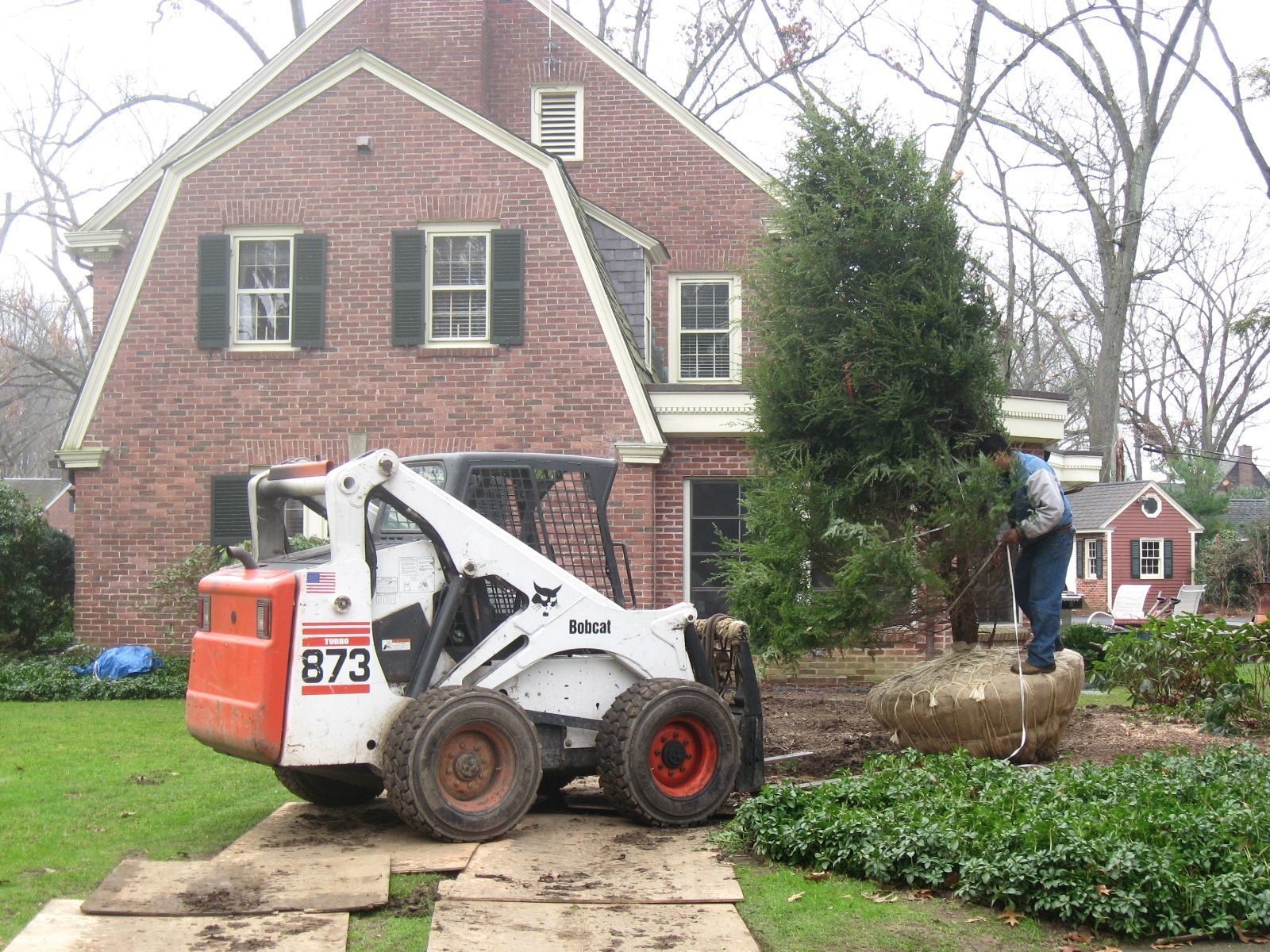 Bobcat operating near a brick house, removing a large stone planter, lawn.