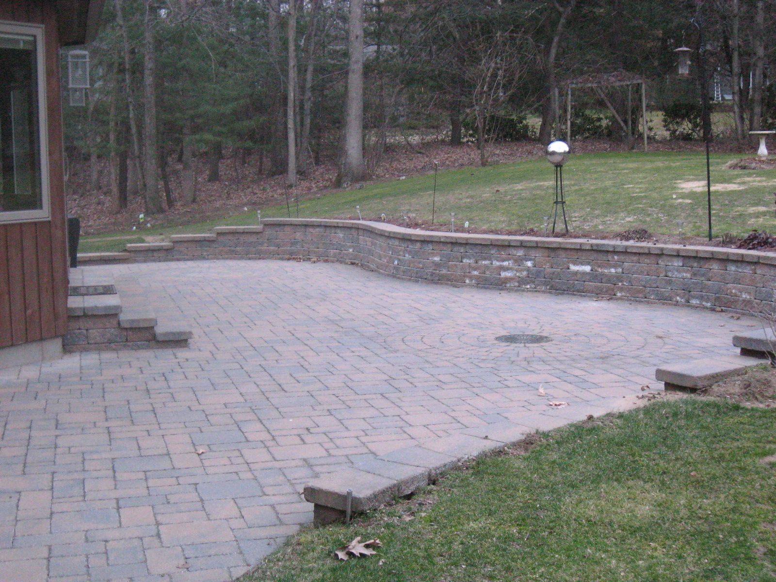 Brick patio with retaining wall, steps leading to wooden house. Green lawn with trees in the background.