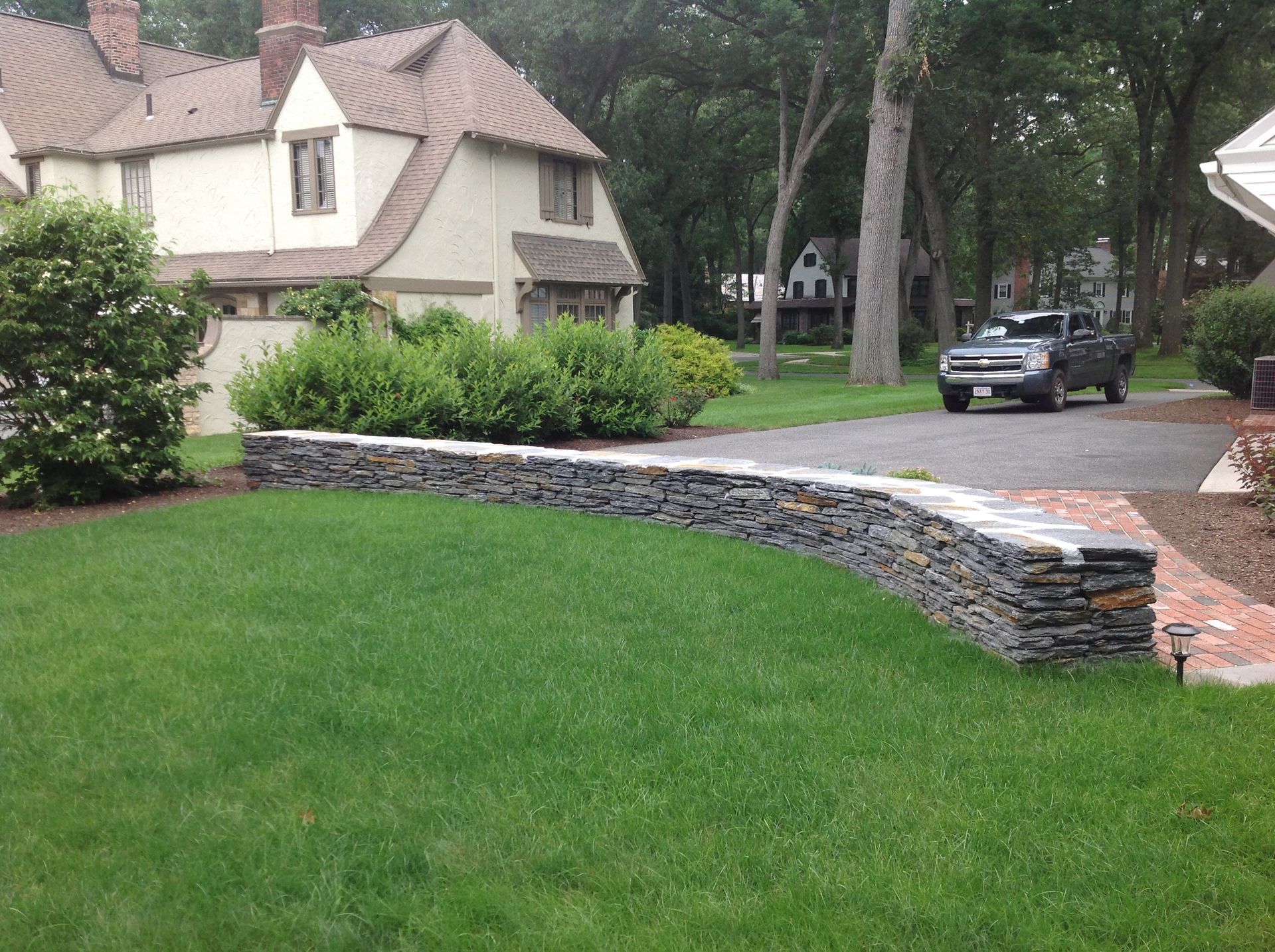 Stone wall borders lush green lawn and driveway; house in background.