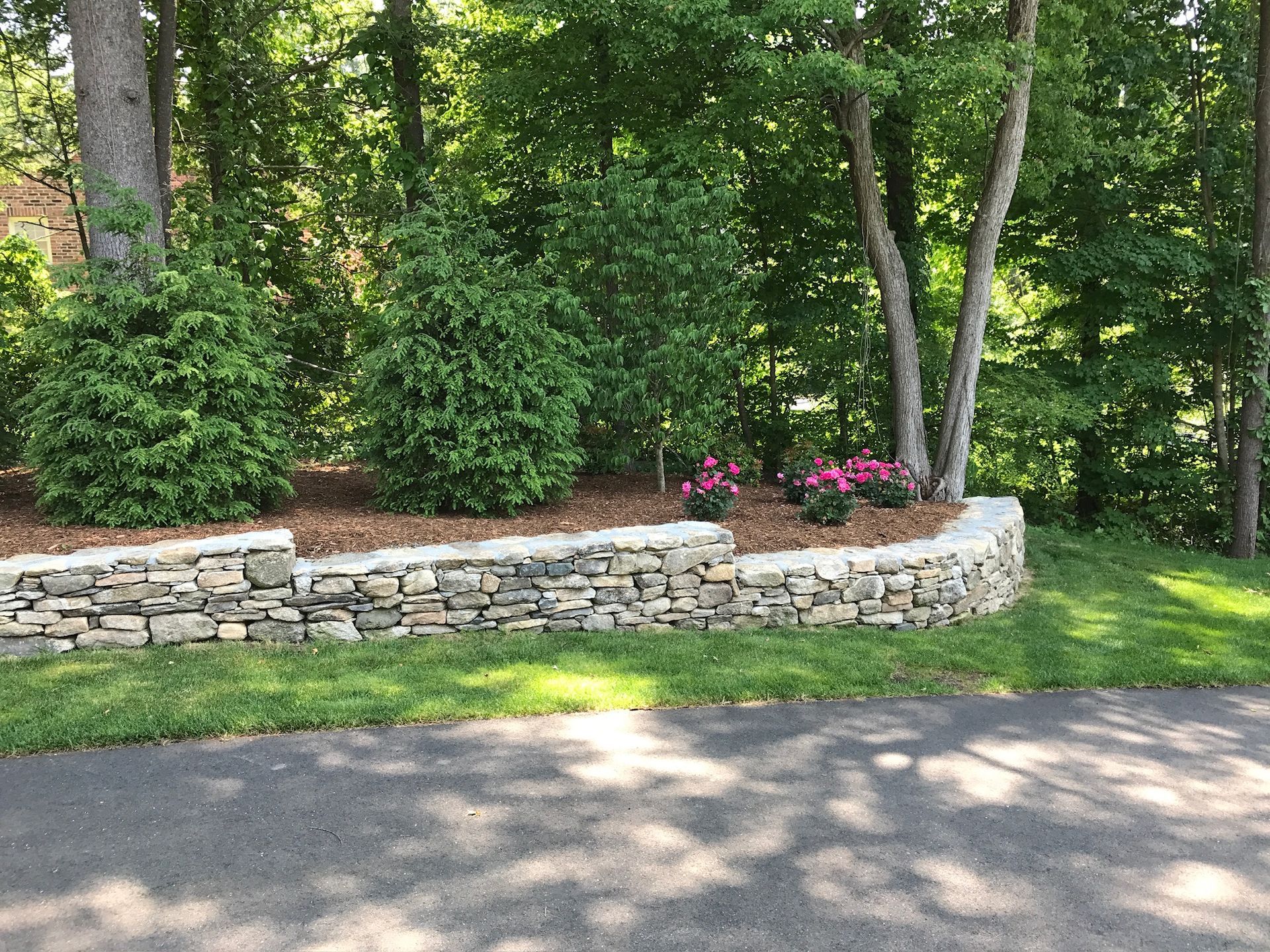 Stone retaining wall with shrubs, mulch, and pink flowers in front of a tree-lined area.
