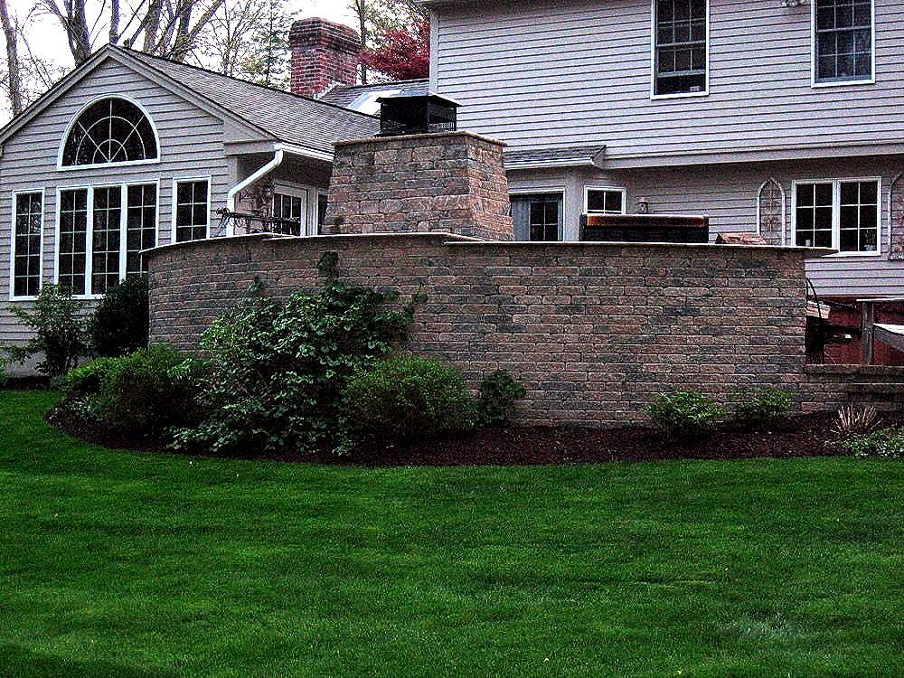 Brick retaining wall with built-in chimney in a yard, with green grass and shrubs against the house.