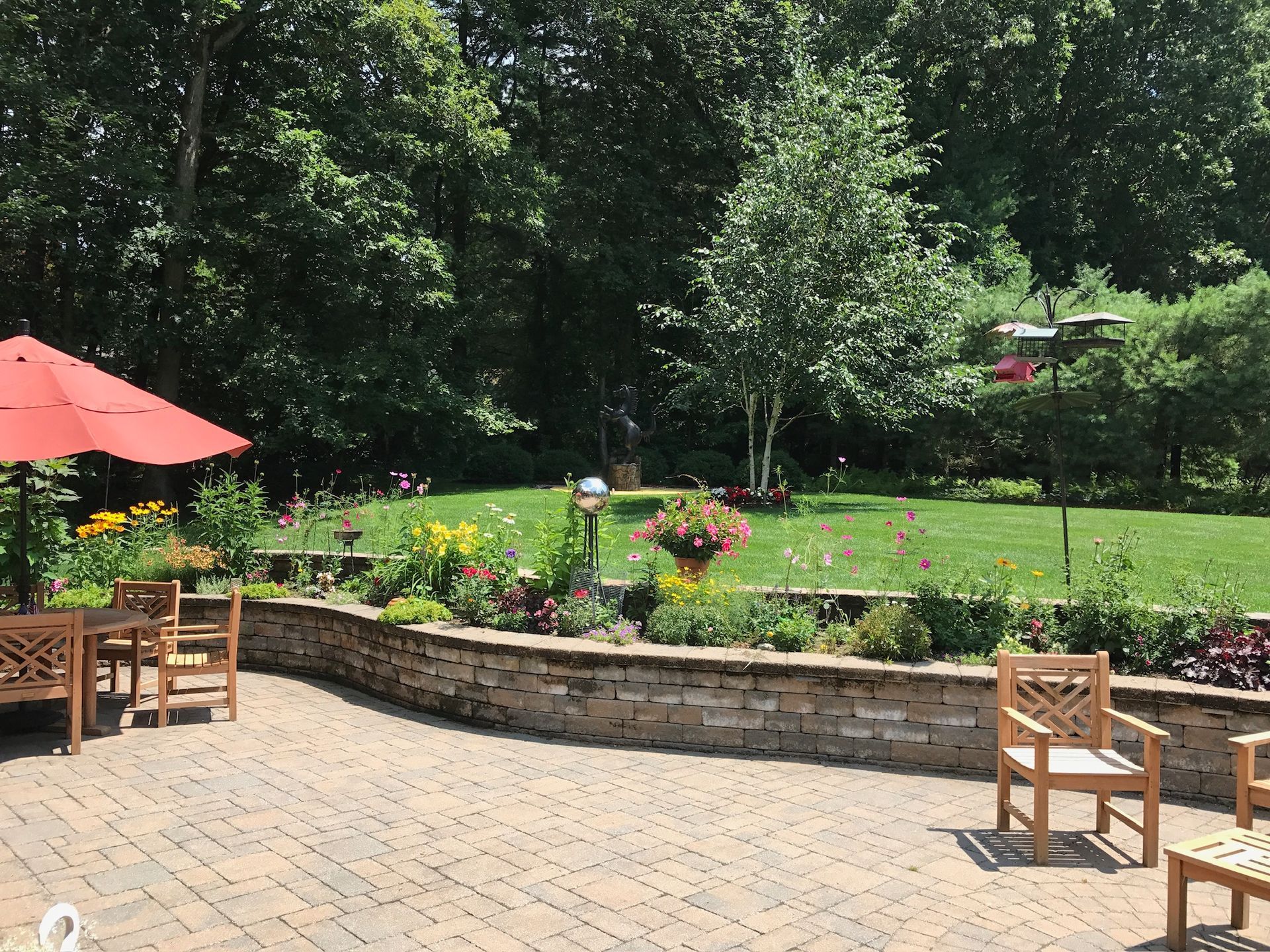 Patio with wooden furniture, red umbrella, flower beds, and a grassy backyard surrounded by trees.