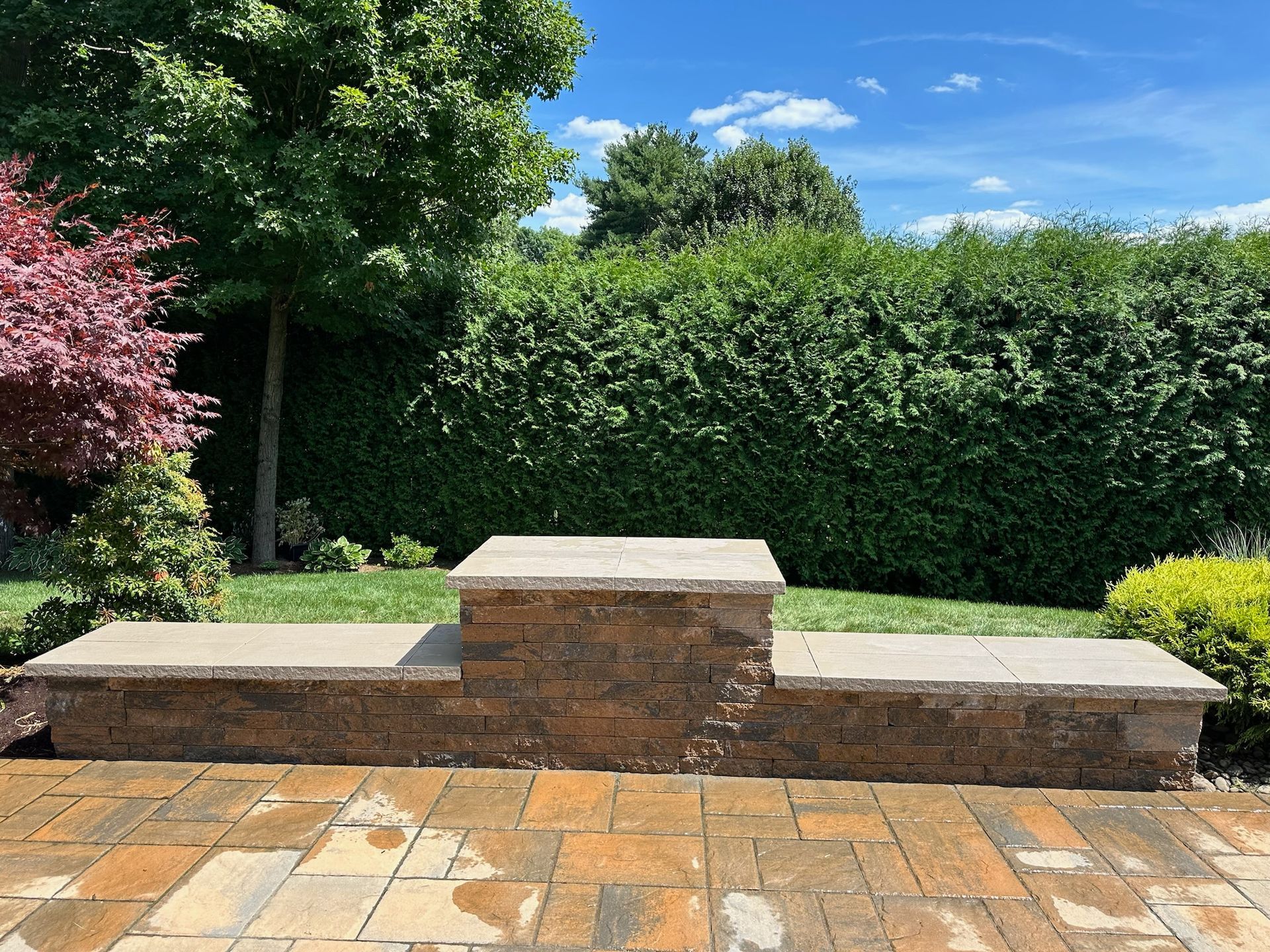 Stone tiered retaining wall in a backyard with lush green hedge and blue sky.