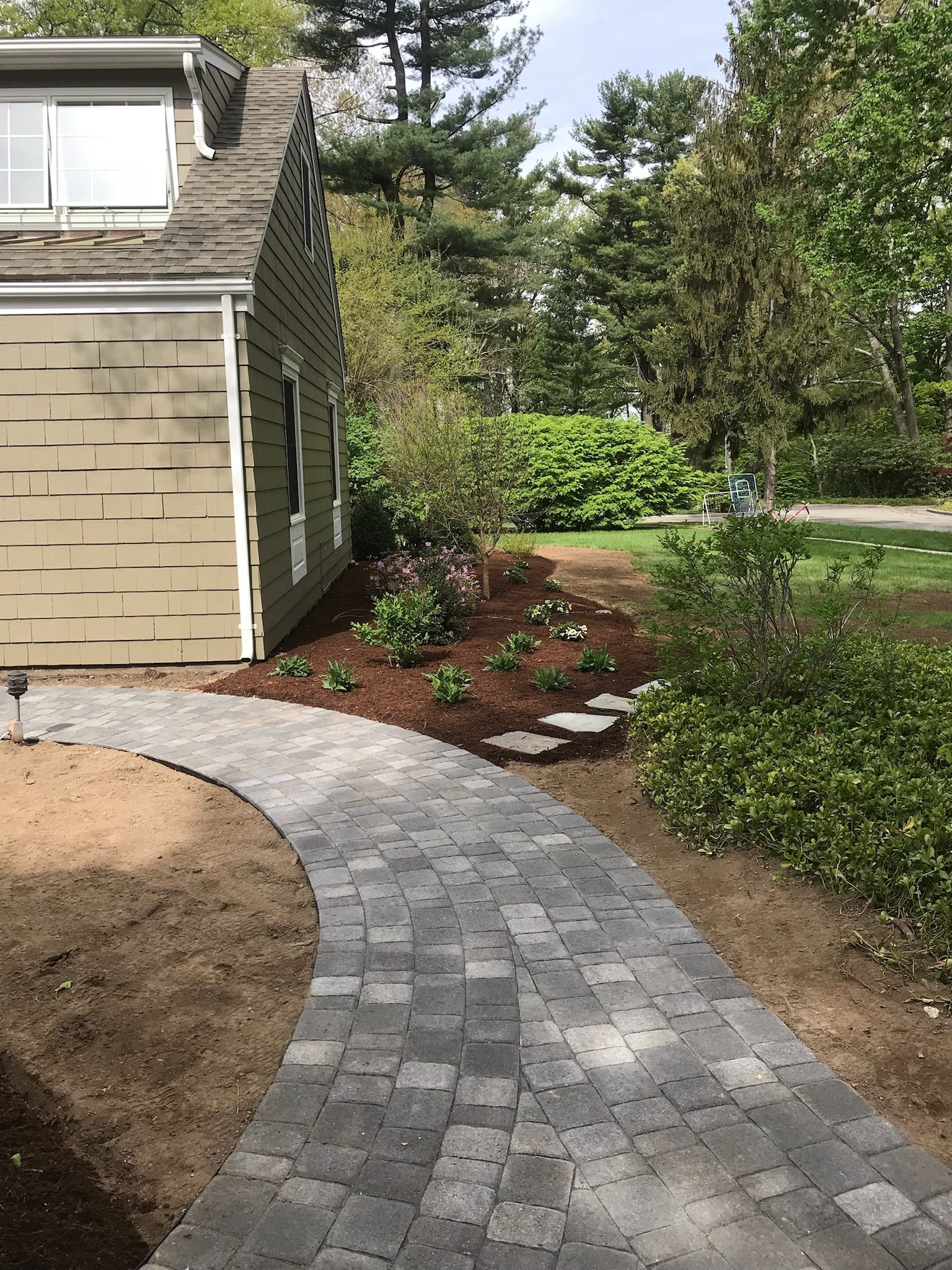 Stone paver walkway curves toward a light green house, with landscaped garden beds.