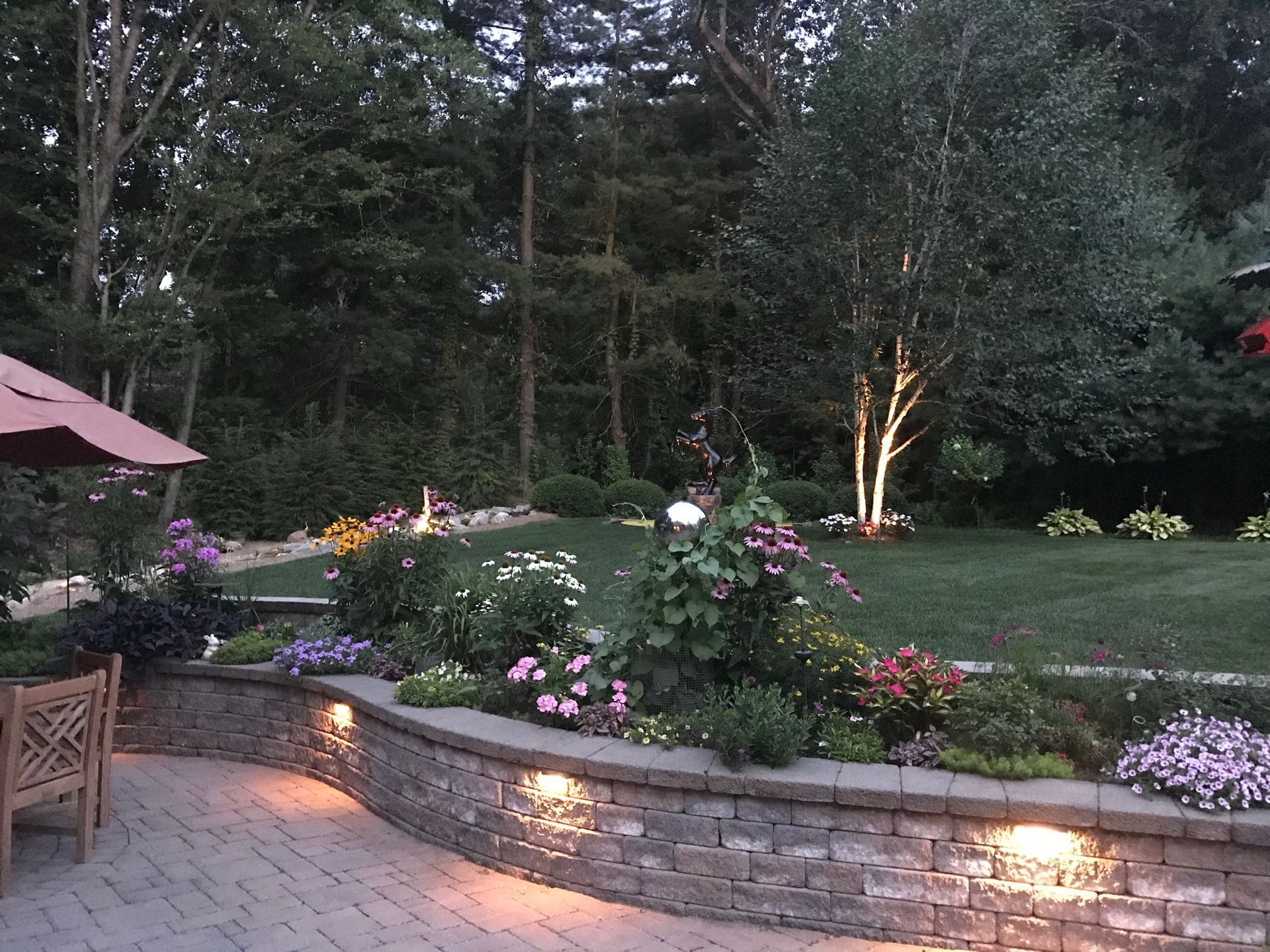 A stone-walled garden bed filled with flowers, with trees in the background, lit by outdoor lights.