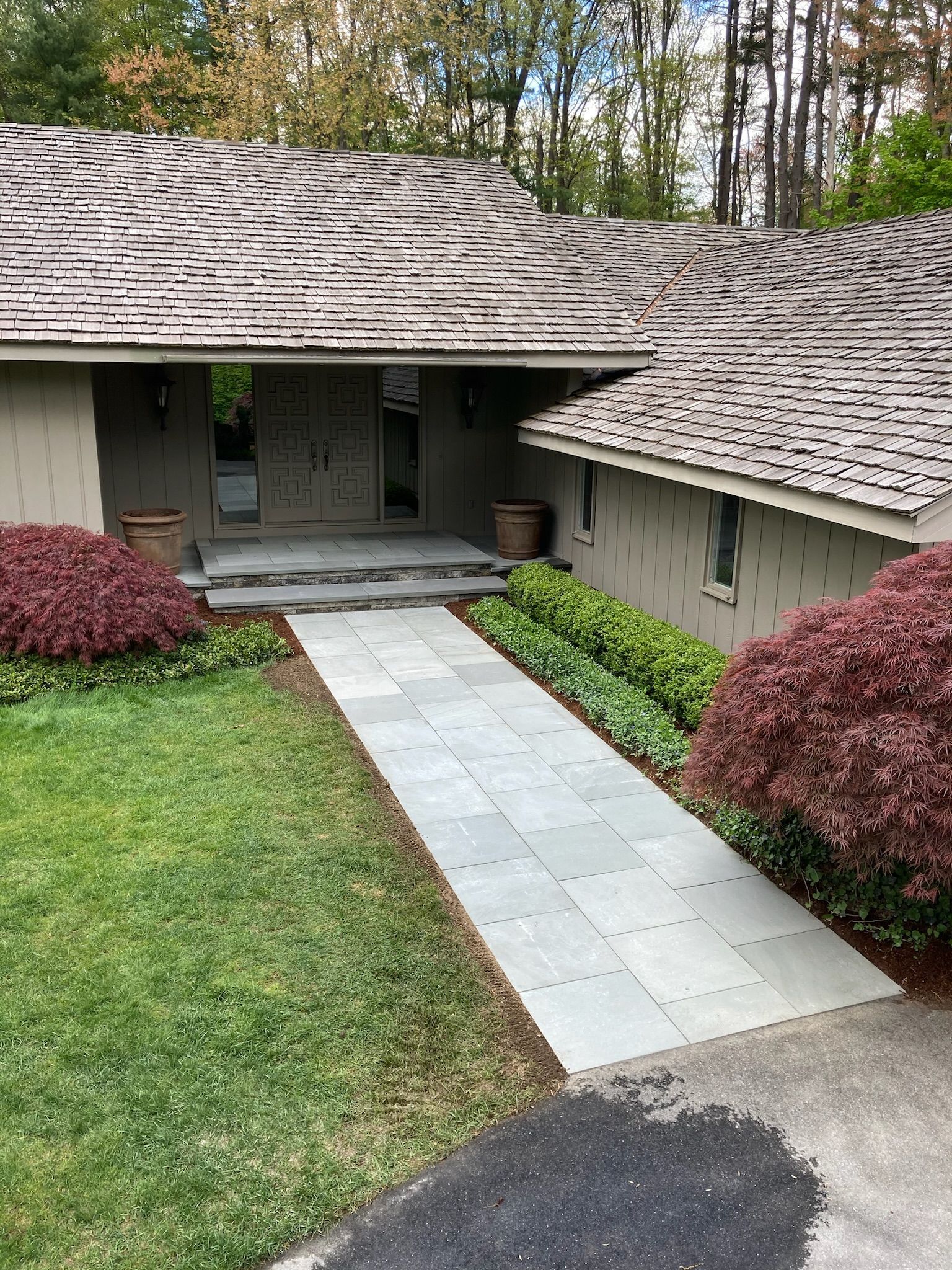 A slate walkway leads to a house with a wooden shingle roof. Landscaping includes red and green bushes.