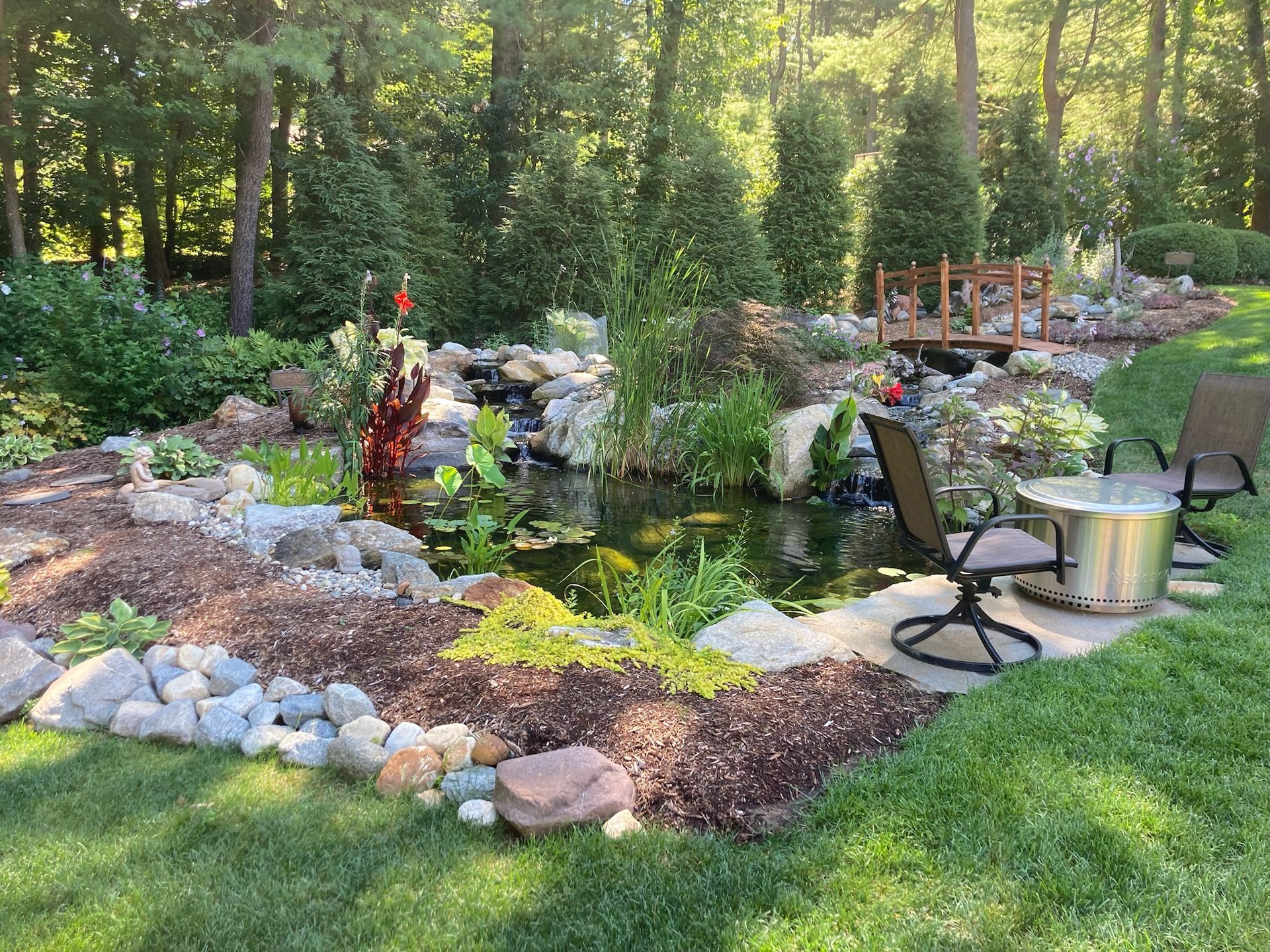 Landscaped backyard with a pond, waterfall, and small wooden bridge. Lawn with seating area.