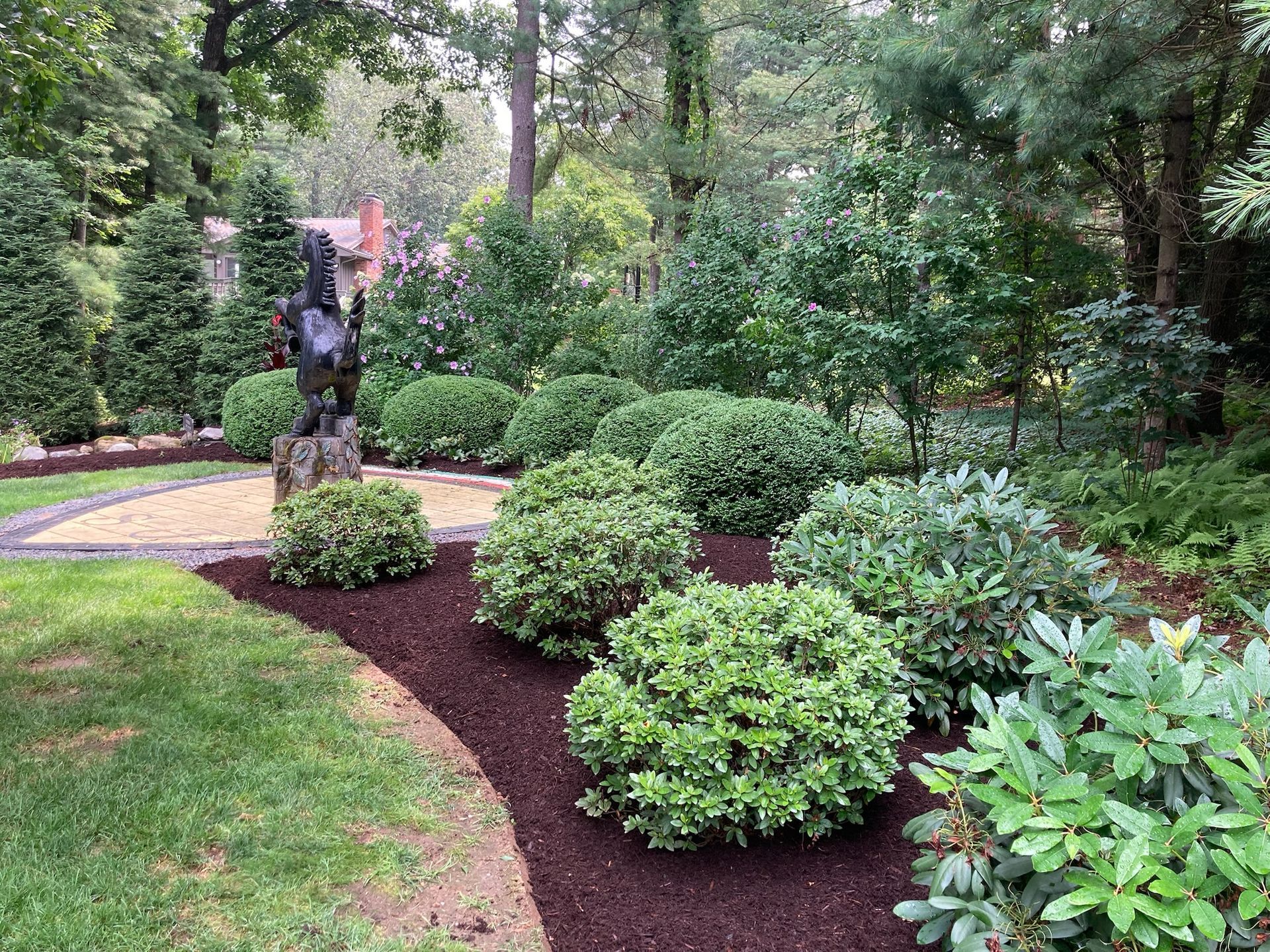 Landscaped garden with mulch, shrubs, and sculpture on a gravel pathway surrounded by greenery.