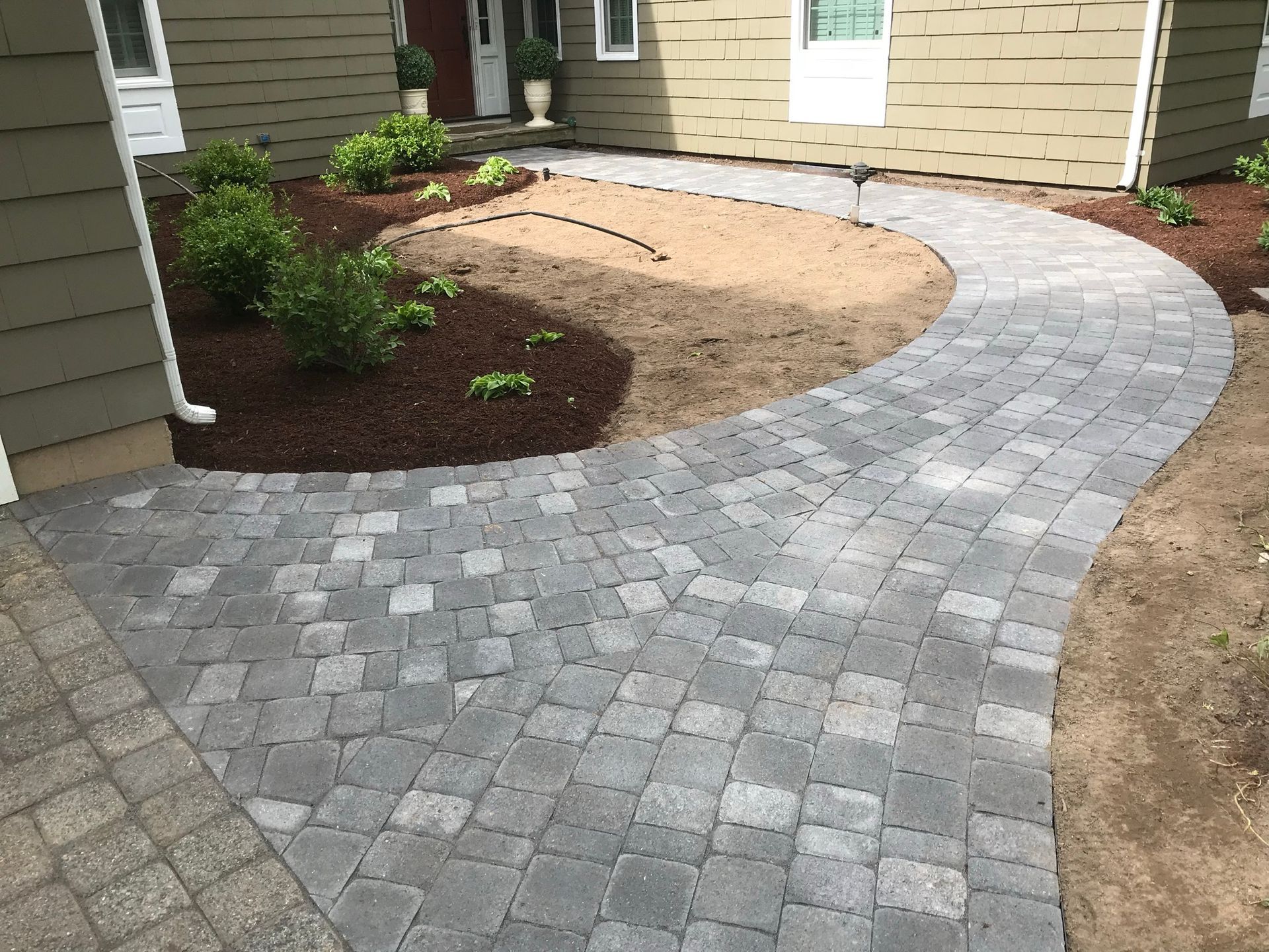Curving brick pathway leading to a house entrance, with mulch landscaping on either side.