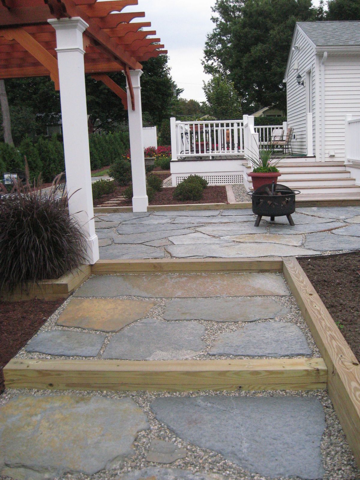 Stone patio with wooden steps leading to a deck and pergola. Blue and tan stone, light wood.