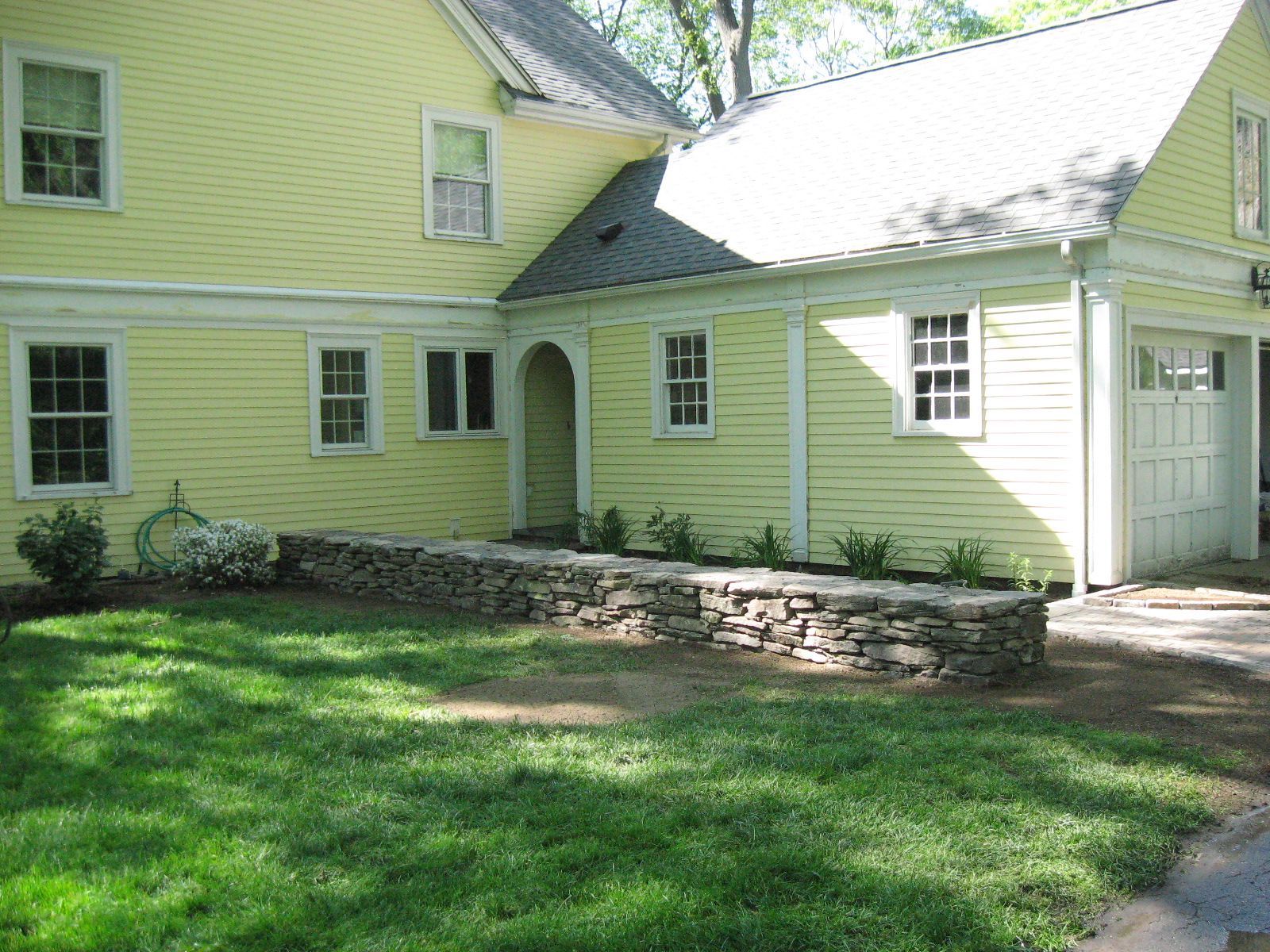 Yellow house with stone wall in front of a green lawn and a driveway.