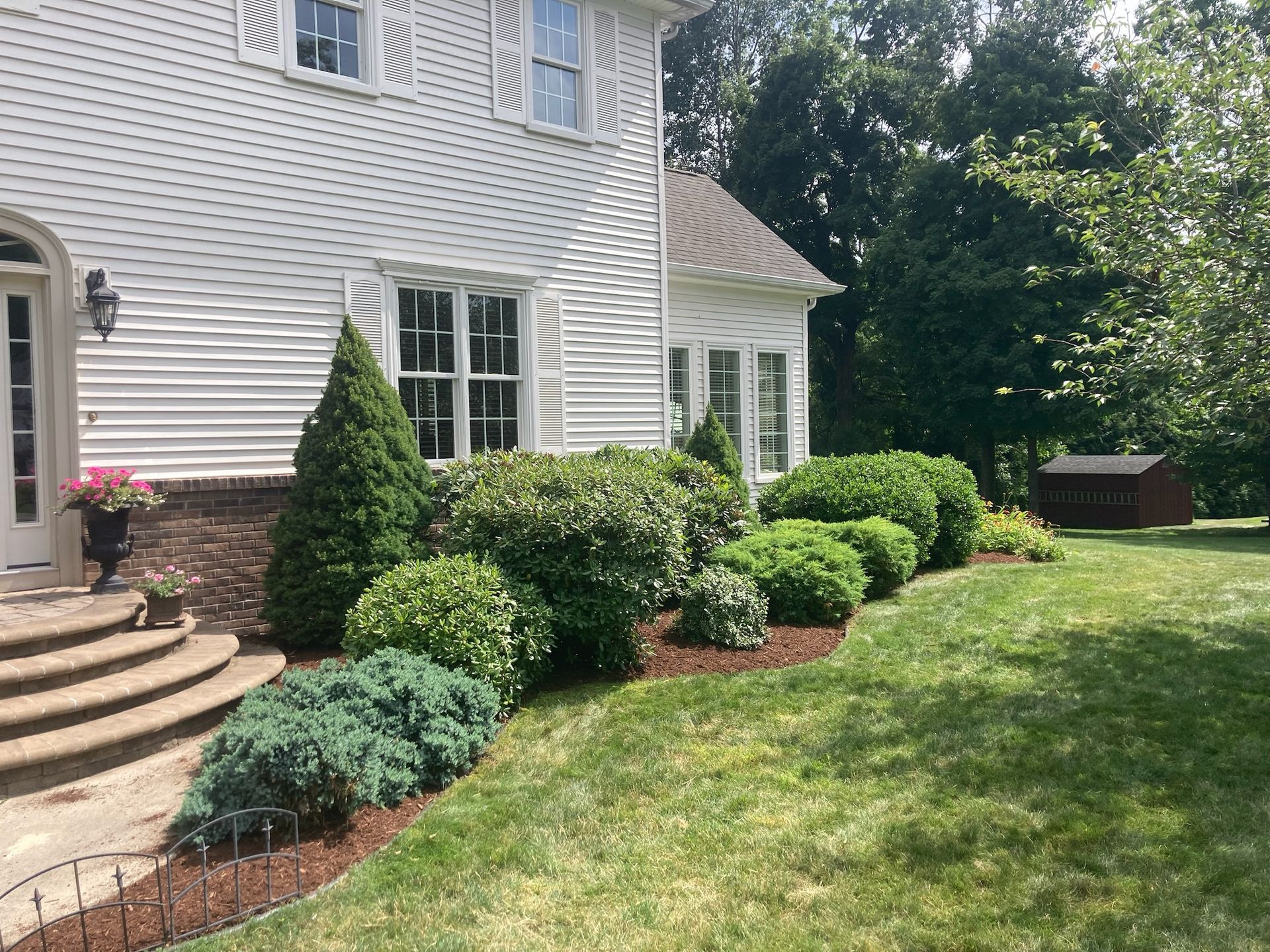 White house exterior with landscaped bed of bushes, trees, and mulch bordering a green lawn.