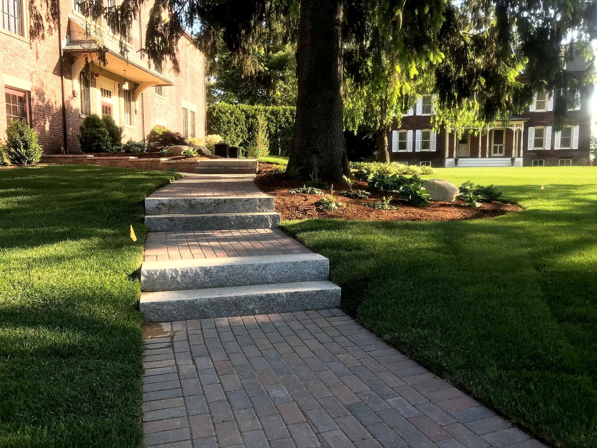 Brick pathway with stone steps leading to a house, surrounded by green grass and trees.