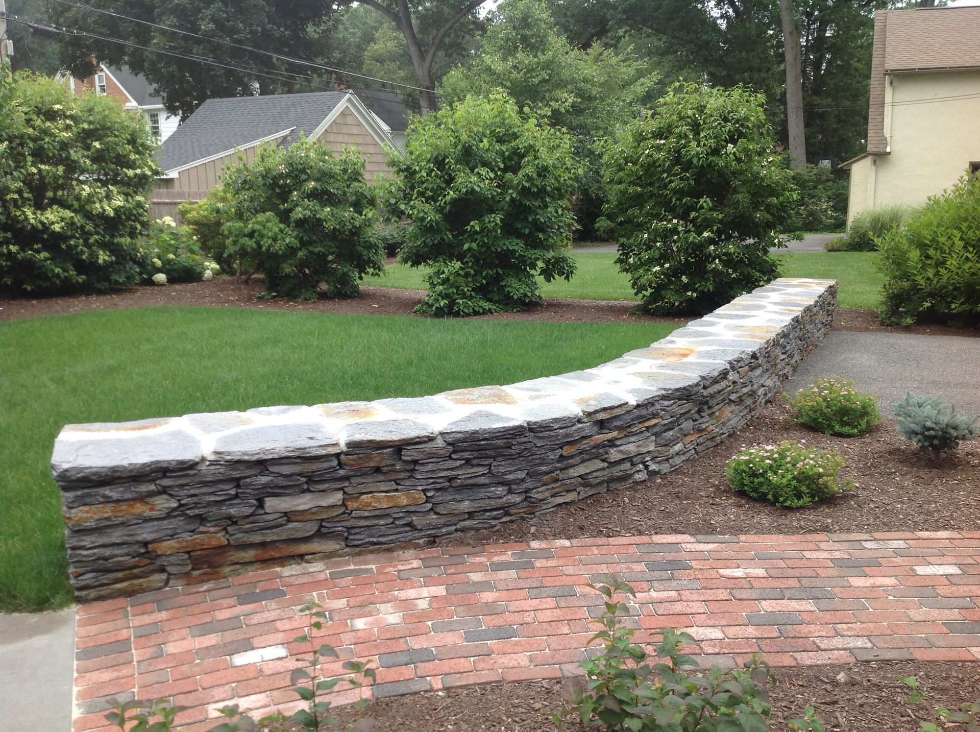 Stone retaining wall curving along a brick pathway and green lawn, with bushes and trees in the background.