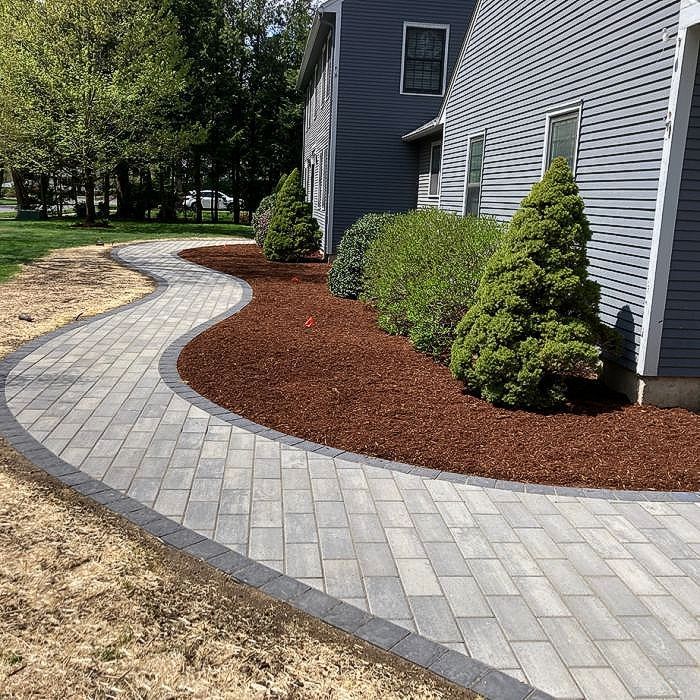 Brick pathway curving along a house with dark siding, brown mulch, and green shrubs.