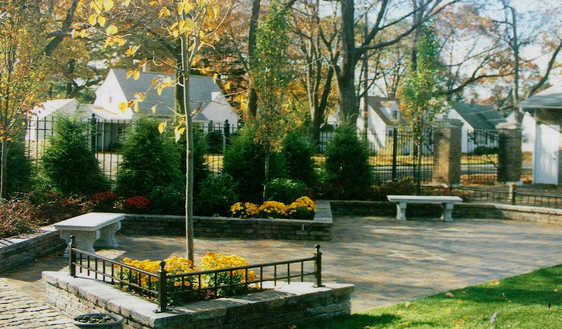 Park with stone benches, trees, and flower beds, bordered by a wrought iron fence and brick walls; houses in the background.