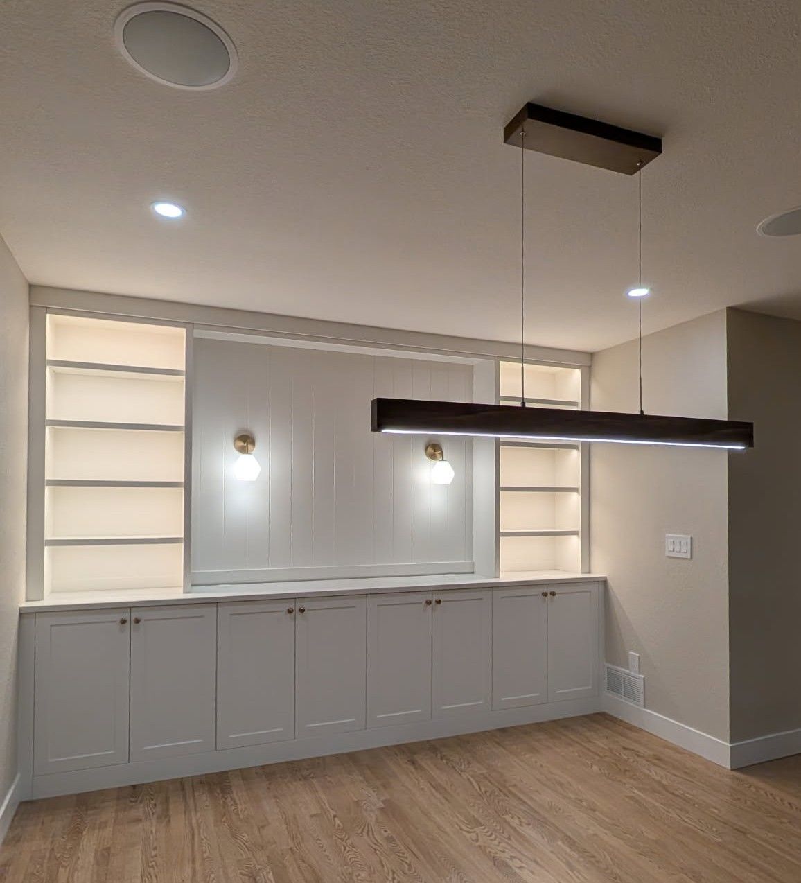 Modern empty dining area with gray cabinets, wood floor, wall sconces, and a hanging linear pendant light.