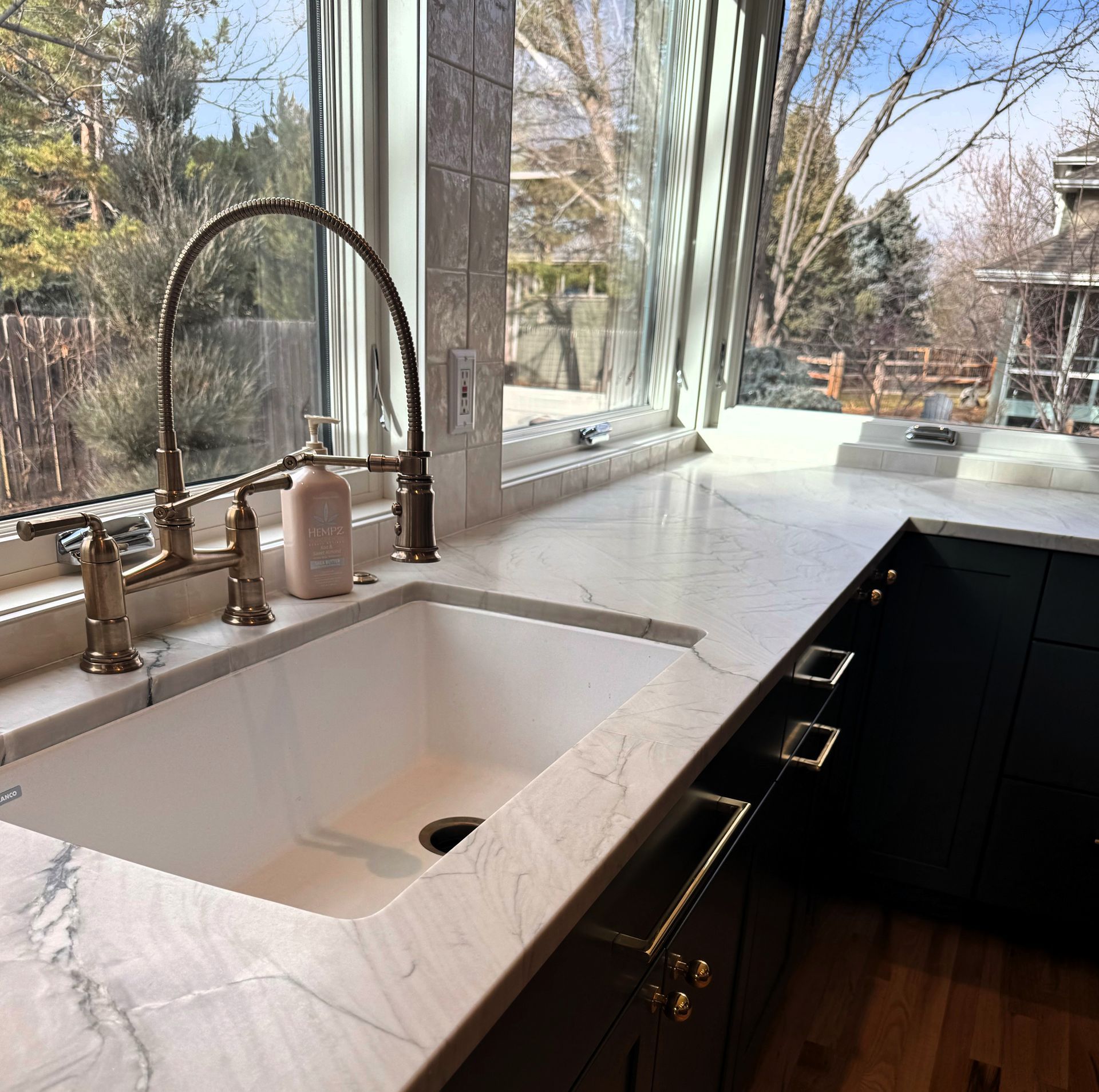 Modern kitchen sink by a large window with white marble countertop, brass faucet, and dark cabinets