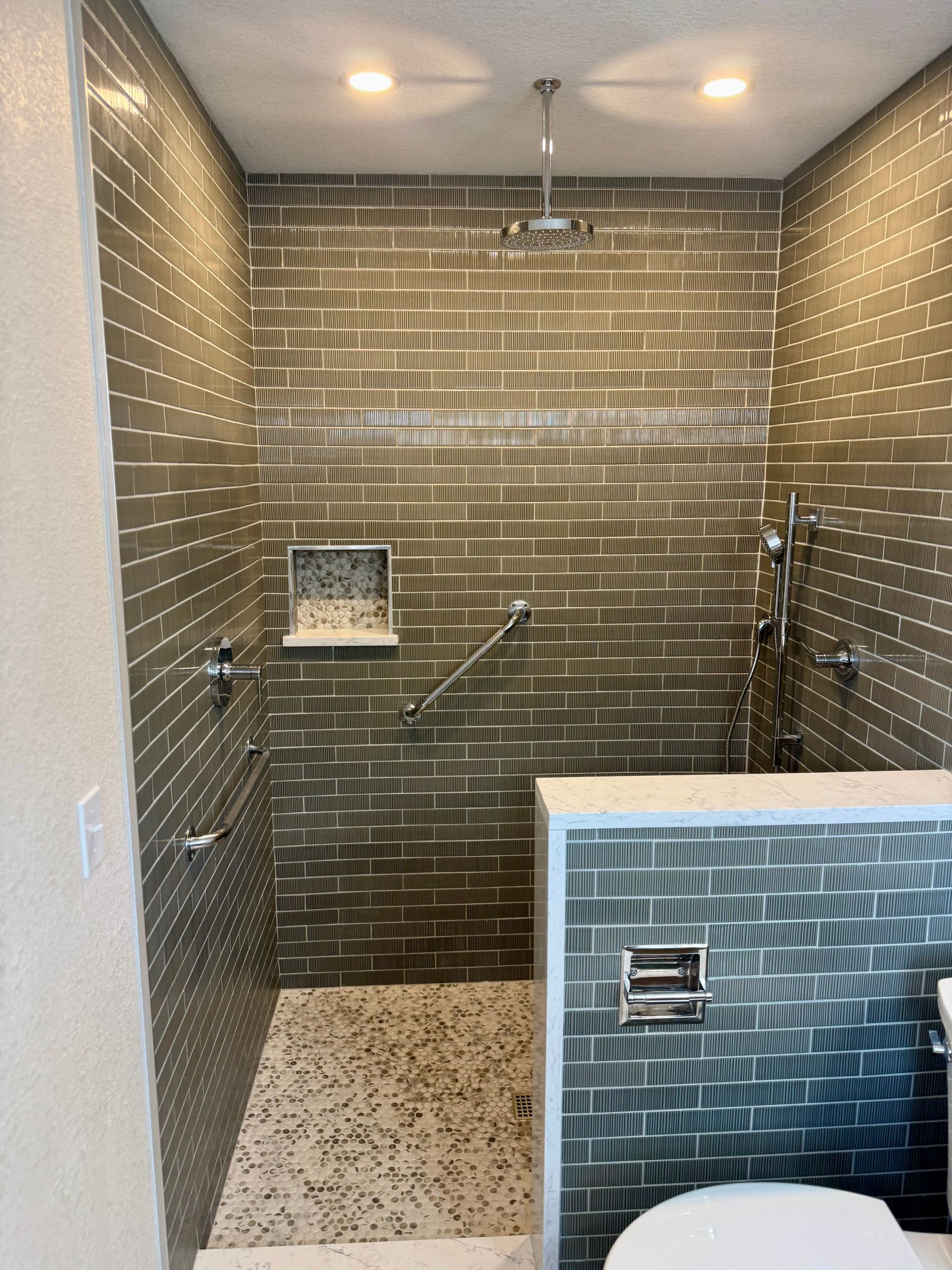 Accessible shower stall with tiled walls, grab bars, bench, and handheld showerhead.