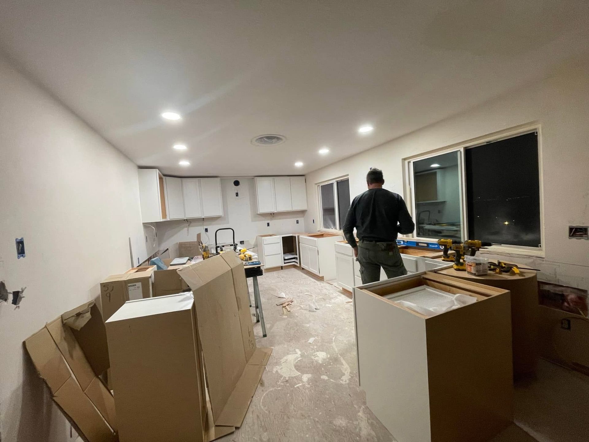 Kitchen under renovation with boxes and debris; a person stands by the counter near a window.