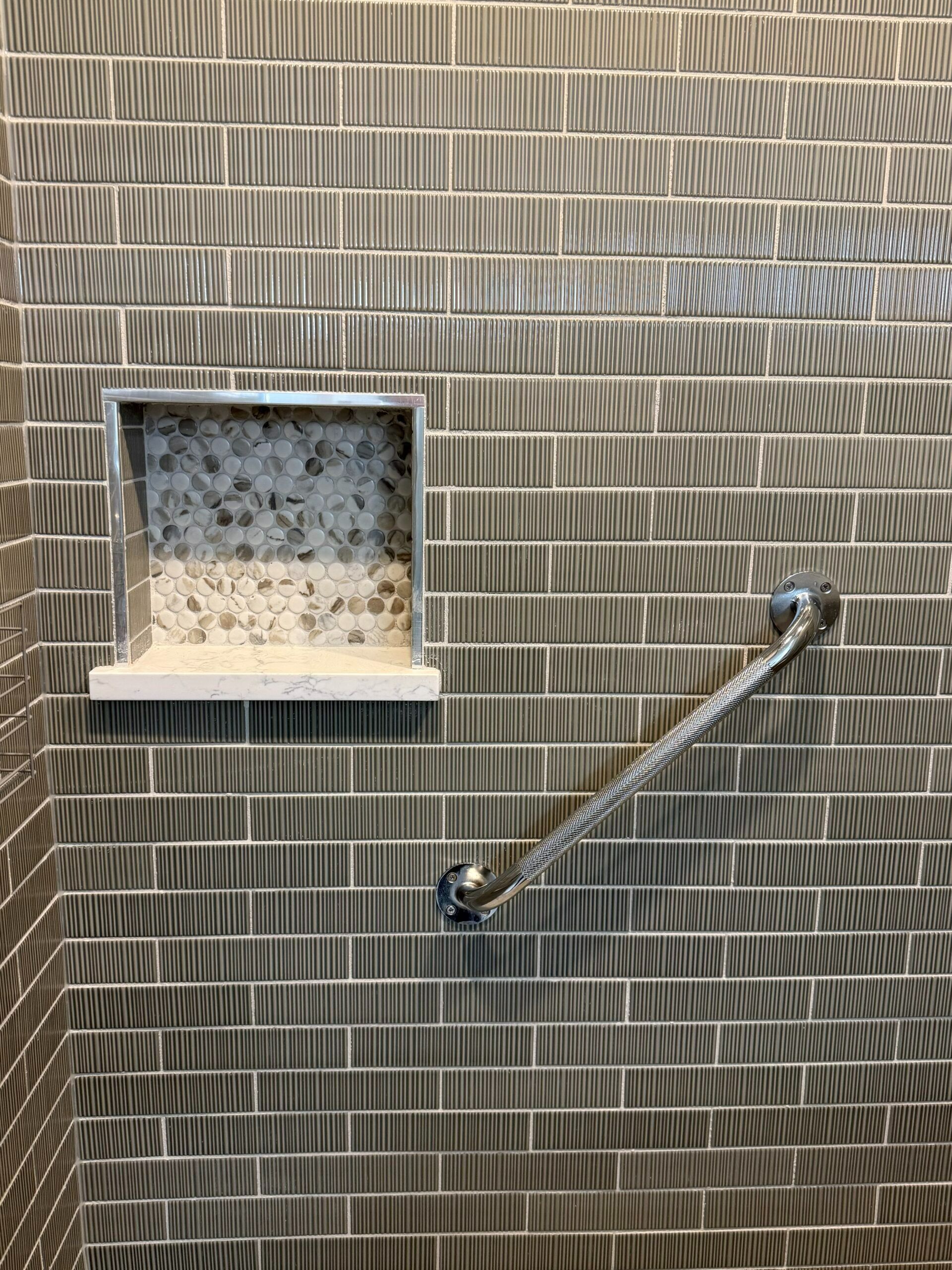 Small square wall niche with pebbles and a ledge, beside a diagonal metal grab bar on textured tiles.