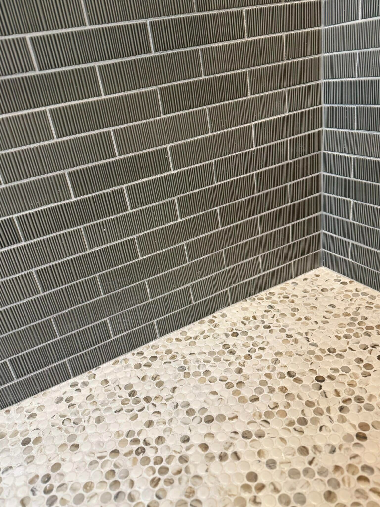 Corner of a tiled wall and pebble-textured floor in a shower or bathroom.