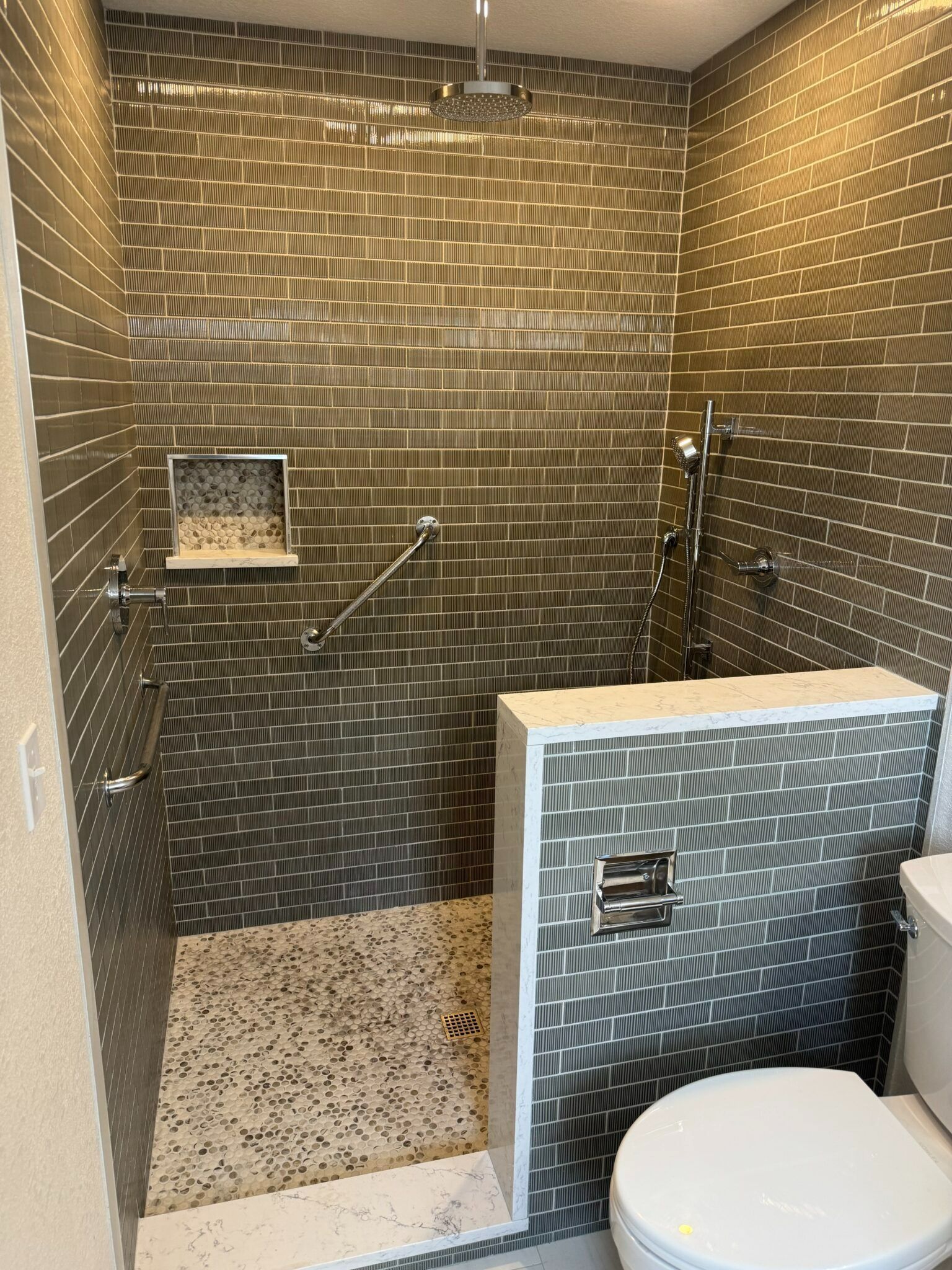 Accessible shower with pebble tile walls, grab bar, corner shelf, and a built-in toilet area in front.