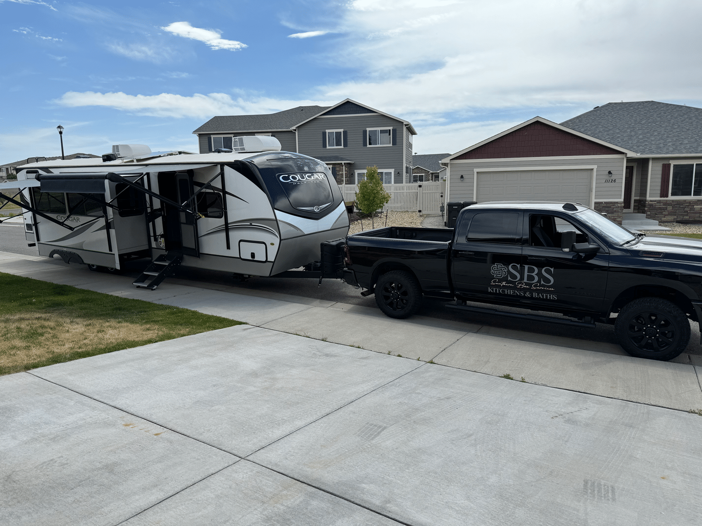Black pickup towing a white travel trailer in a suburban driveway with houses in the background
