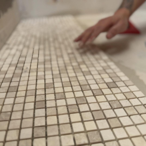 Hand placing white mosaic tiles on a tiled floor in a room under construction