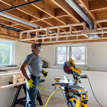 Worker standing in a room under renovation beside a miter saw and exposed ceiling framing.