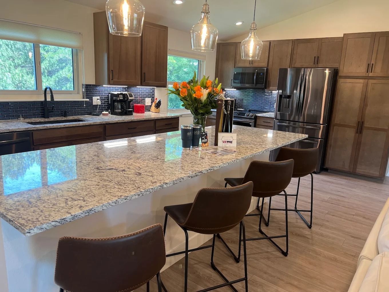 Modern kitchen with large granite island, brown bar stools, stainless fridge, and pendant lights