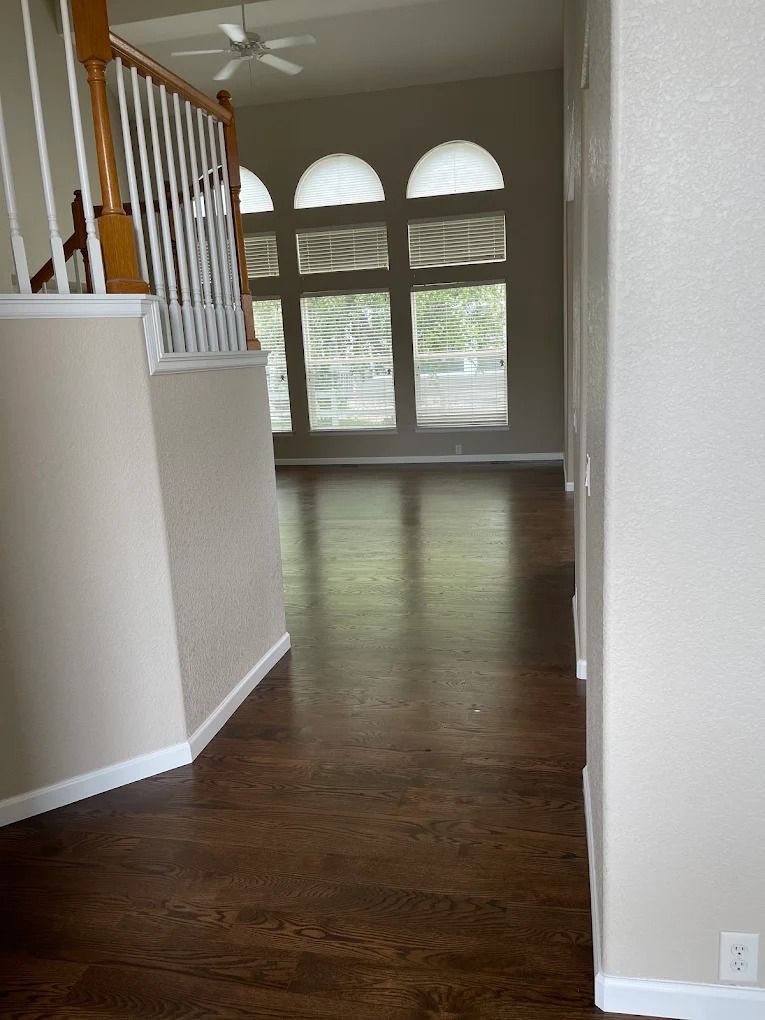 Empty hallway with dark wood floors, white walls, staircase, and large arched windows at the far end