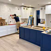 Person painting a wall in a bright kitchen with white cabinets and a blue island under renovation