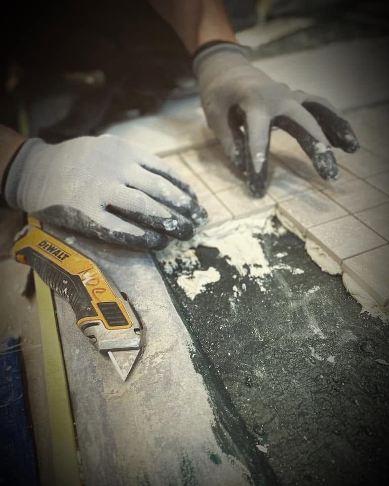 Gloved hands smoothing dark sealant on tiled floor with a utility knife nearby