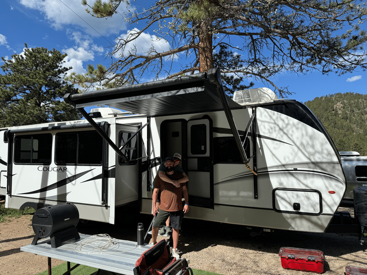 Person standing at the doorway of a white camper trailer under tall pine trees on a sunny day