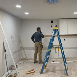 Worker on a ladder in a kitchen under renovation, with tools and cabinets visible.