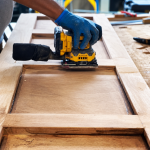 Gloved hand using a yellow sander on a wooden cabinet door in a workshop