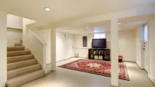 Bright basement living room with beige carpet, red area rug, TV, and stairs on the left