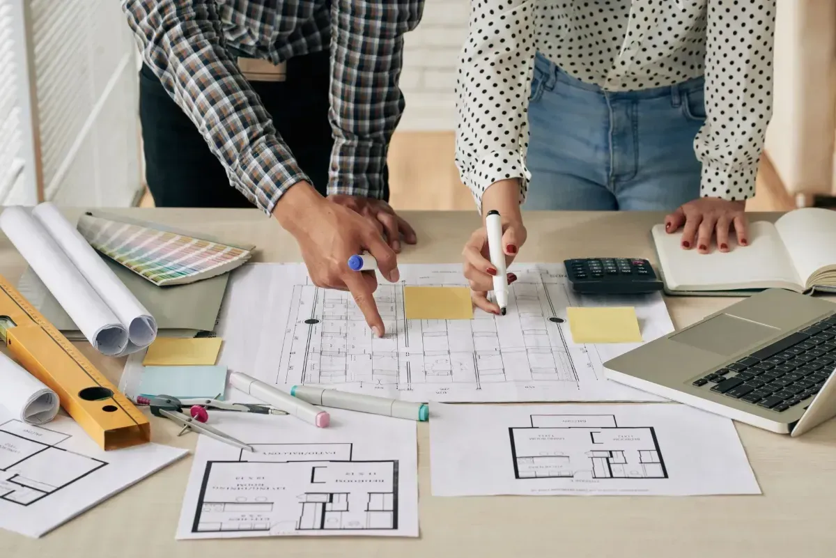 Two people reviewing floor plans and blueprints at a desk with laptop, ruler, and pencil.