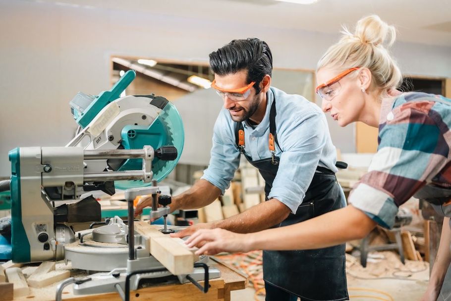 Two people using a miter saw in a workshop, focused on cutting wood boards.