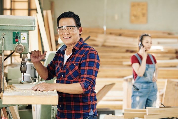 Man smiling beside a drill press in a woodshop, with a woman working in the background